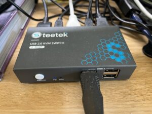 A photo of the Steetek KVM switch, with a USB cable plugged in the front and various USB and HDMI cables plugged into the back. It's a grey, rectangular metal box.